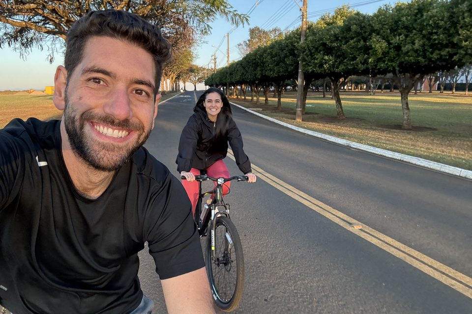 Renan em primeiro plano e Ana pedalando atrás, numa avenida arborizada